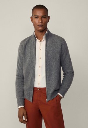 Cardigan grigio con zip in morbido materiale knit, caratterizzato da polsini a coste e una vestibilità moderna, abbinato a una leggera camicia con bottoni e pantaloni color ruggine.