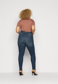 Krulligt hårig kvinna med en brun kortärmad topp, blå högmidjade skinny jeans och svarta högklackade sandaler, står mot en vit vägg.