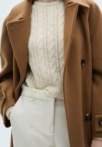 Beiger Wollmantel mit großen Revers und schwarzen Knöpfen, über einem cremefarbenen Kabelstrickpullover, kombiniert mit weißen Hosen. Sanfte Texturen und Lagenlook.
