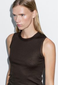 Brązowy bezrękawnik o dopasowanym kroju, wykonany z lekkiego materiału, z okrągłym dekoltem i lekko przeźroczystymi akcentami.