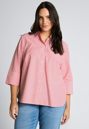 Femme portant une chemise à rayures verticales roses et blanches avec des manches 3/4 et un jean bleu, debout devant un arrière-plan uni.