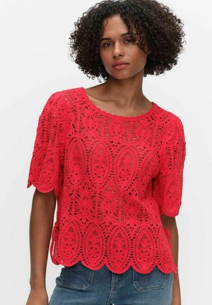 Kobieta ubrana w czerwony krótki top z szydełkowym wzorem kwiatowym i falbankowym brzegiem, połączony z niebieskimi dżinsami na jednolitym tle.