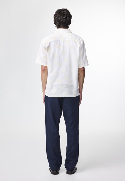 Korte mouwen, witte blouse met een gestructureerd schelpatroon, gecombineerd met losse marineblauwe broek en zwarte schoenen. Eenvoudig knoopsluiting ontwerp.