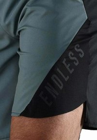 Sportshorts mit einem grünen oberen Abschnitt und einem schwarzen unteren Abschnitt, versehen mit einem vertikalen Text "ENDLESS" in einem strukturierten Muster an der Seite.