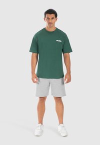 Camiseta de algodón verde con escote redondo y mangas cortas, que presenta el texto "SMILODOX" en blanco. Combinada con pantalones cortos grises y zapatillas blancas.