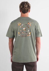 T-shirt en coton vert olive avec un motif floral coloré dans le dos, à manches courtes et col rond. Logo Oxbow inclus.