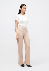 Biela priliehavá tričko s rebrovanou textúrou; svetloružové široké nohavice s jemným vzorom; čierne lodičky so špicatou špičkou. Modelka stojí v profile.