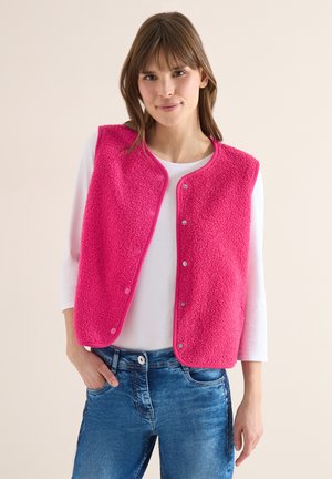Femme portant un gilet polaire rose vif sur un chemisier blanc à manches longues et un jean bleu, debout avec une main dans la poche devant un fond uni.