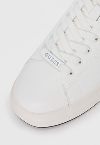 Baskets blanches avec une tige en cuir lisse, bout arrondi, fermeture à lacets plate et une petite étiquette bleue affichant la marque "GUESS."