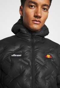 Schwarze gepolsterte Jacke mit hohem Kragen, ausgestattet mit einem Reißverschluss, strukturiertem Stoff und einem roten, gelben und schwarzen Logo auf der Brust.