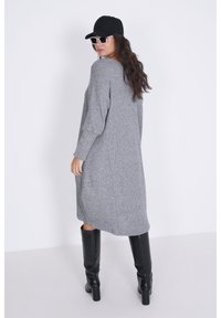 Grijze gebreide jurk met lange mouwen, een ontspannen silhouet en zijzakken, gecombineerd met zwarte kniehoge laarzen en een zwarte pet.