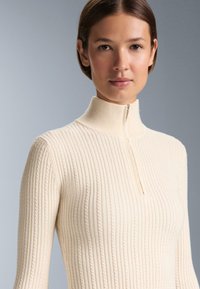 Kremasti rebrasti pleteni top s visokim izrezom i polu-zip zatvaranjem. Ima teksturirane linije kroz cijeli materijal, naglašavajući njegov prilagođeni dizajn.