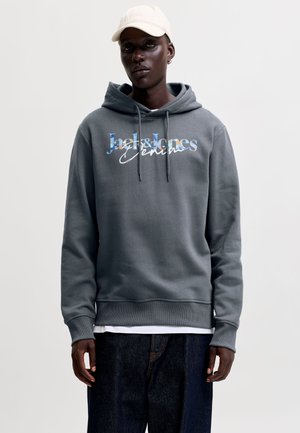 Joven con sudadera gris con capucha con texto "Jack & Jones Denim", vaqueros oscuros, gorra blanca y un pendiente, de pie delante de un fondo liso.