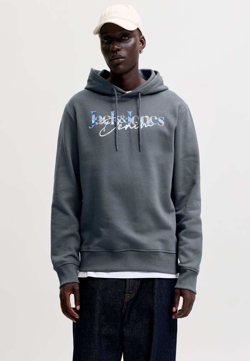 Jeune homme portant un sweat à capuche gris avec le texte "Jack & Jones Denim", un jean foncé, une casquette blanche et une boucle d’oreille, debout devant un fond uni.