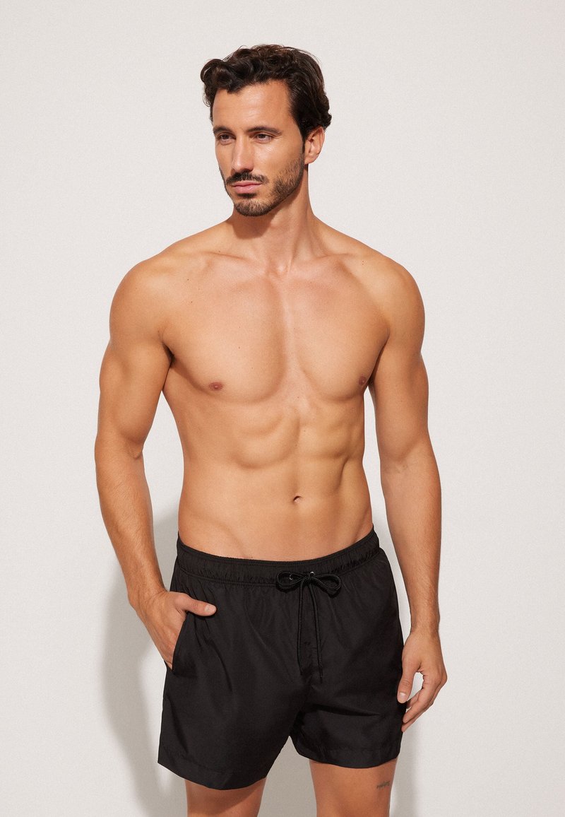 Muskelbepackter Mann mit kurzen dunklen Haaren, der schwarze Badeshorts trägt, steht mit einer Hand in der Tasche und schaut seitlich vor einem schlichten Hintergrund.