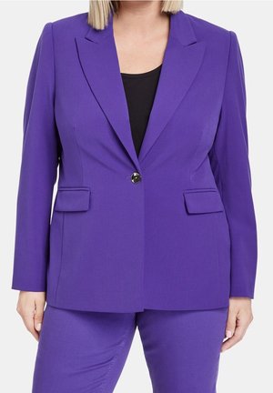 Vrouw die een op maat gemaakt paarse blazer draagt met een enkele zwarte knoop over een zwarte top, gecombineerd met bijpassende paarse broek.