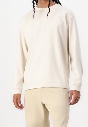 Camicia beige a maniche lunghe realizzata in tessuto morbido, con scollatura a girocollo e polsini a coste, abbinata a pantaloni beige chiaro.