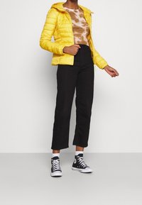 Gul pufferjakke med hætte, tekstureret overflade, parret med en brun tie-dye skjorte og sorte cropped bukser; stylet med sorte high-top sneakers.