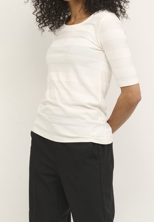 Femme aux cheveux bouclés portant un haut blanc texturé à manches courtes et un pantalon noir, posant avec les mains dans le dos devant un fond uni.