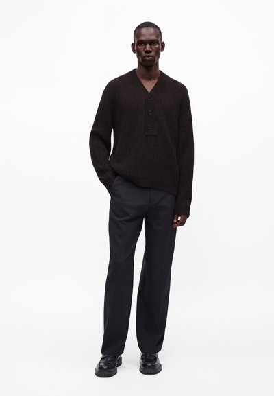 Homme portant un pull côtelé foncé avec des boutons, un pantalon noir et des chaussures noires, debout devant un fond blanc uni.
