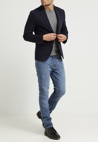 Navy blazer over a grey shirt, paired with light blue distressed jeans and black shoes. Features structured fit and classic design elements.