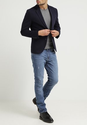 Navy blazer over a grey shirt, paired with light blue distressed jeans and black shoes. Features structured fit and classic design elements.