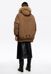 Chaqueta acolchada marrón con capucha de piel sintética, mangas largas y dobladillo elástico, combinada con una falda de cuero negra brillante y botas hasta la rodilla.