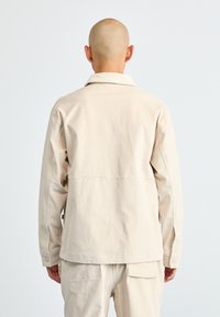 Persone con testa rasata che indossa una camicia a maniche lunghe beige e pantaloni abbinati, mostrata da dietro su uno sfondo bianco semplice.