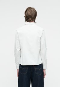 Camicia bianca a maniche lunghe con un retro fitted e colletto classico, abbinata a pantaloni in denim scuri con cuciture visibili e tasche.