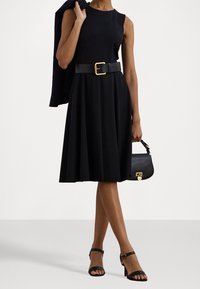 Svart A-linje klänning med ärmlös design, åtsittande midjebälte och utsvängd kjol. Tillsammans med en svart handväska och klackskor med remmar.