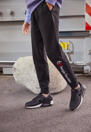 Person iført sorte Sergio Tacchini joggingbukser og sorte Nike Air Max sneakers står på grå asfalt ved siden af en stor sten.