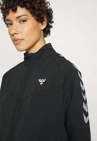 Nahaufnahme einer Person, die eine schwarze Jacke mit Reißverschluss mit weißen Chevron-Streifen am Ärmel und einem kleinen weißen Bienenlogo auf der Brust trägt.