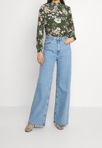 Grön blus med blommönster och långa ärmar, hög hals, i kombination med ljusblå vida jeans. Modellen står med en hand på höften och bär blå sandaler.