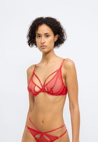 Rød gjennomsiktig mesh lingerie-sett som har en BH med trekantet design, kryssende stropper og dekorative metallbeslag.