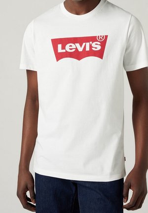 Mężczyzna w białej koszulce Levi's z czerwonym logo i ciemnoniebieskich jeansach, stojący na jednolitym jasnym tle.