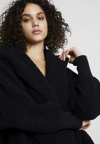 Veste en polaire noire avec une surface texturée, large col châle et poignets côtelés, offrant une coupe décontractée et des manches longues.