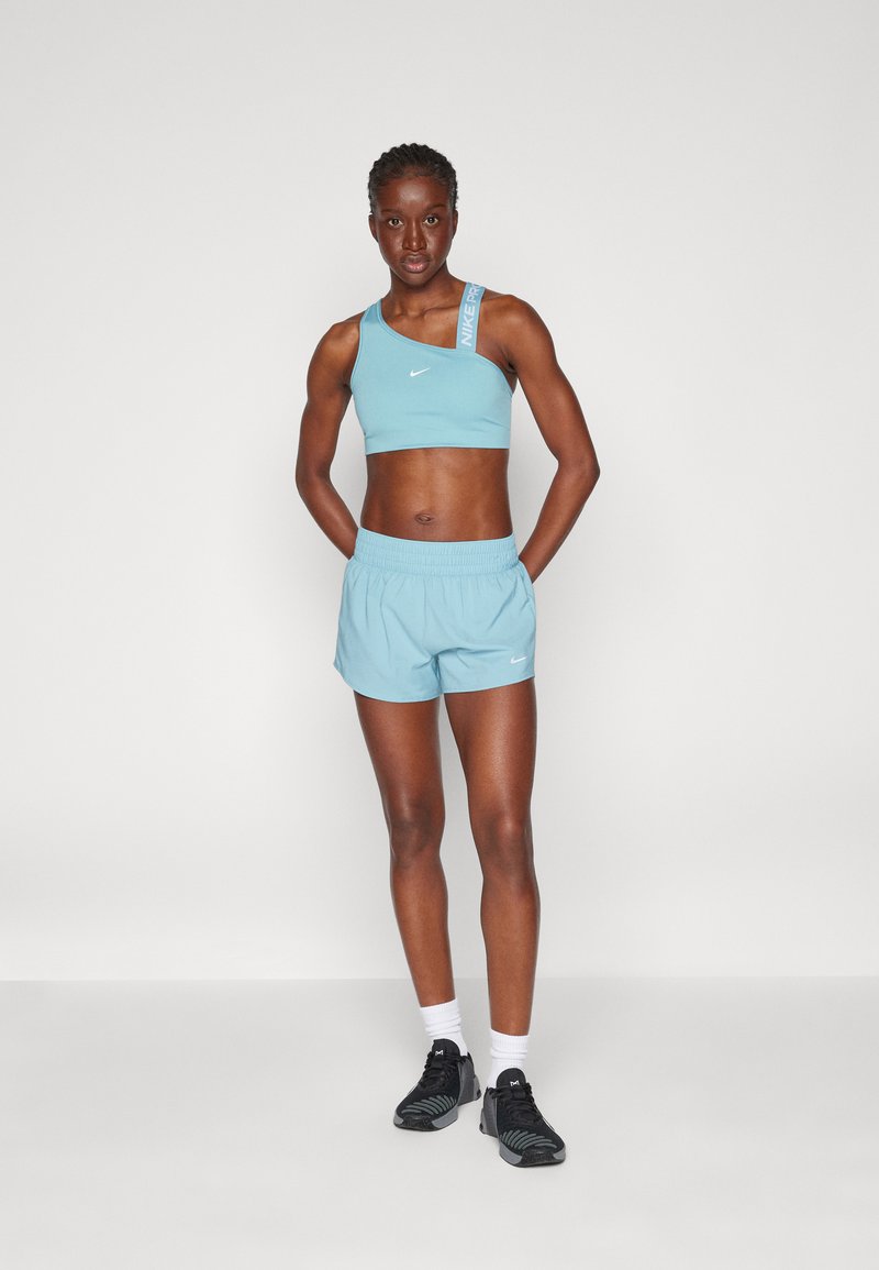 Hellblaue Sport-BH mit einseitigem Design und weißem Logo, kombiniert mit passenden hellblauen Shorts. Schwarze Sportschuhe mit weißen Socken.