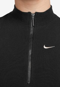 Černý zip-up top s vysokým límcem vyrobený z ribbrované tkaniny. Na hrudi se nachází kontrastní bílé logo Nike a hladký zip.