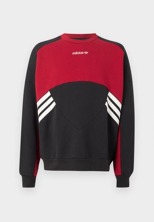 Ung mand med krøllet hår bærer en sort kasket bagvendt, en rød og sort Adidas sweatshirt og lyseblå løstsiddende jeans mod en ensfarvet baggrund.