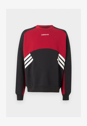 Fiatal férfi göndör hajjal, fekete hátrafordított sapkában, piros-fekete Adidas pulóverben és világoskék, bő fazonú farmernadrágban, sima háttér előtt.