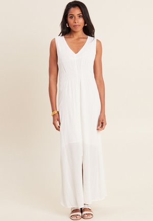 Witte mouwloze maxi-jurk met een V-hals, gestructureerde stof en een split aan de voorkant. Model draagt bruine sandalen en gouden creolen.