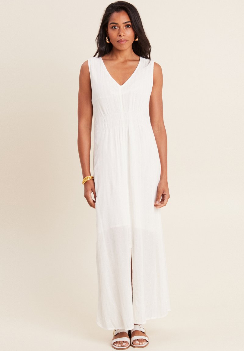 Robe maxi sans manches blanche avec un décolleté en V, en tissu texturé, et une fente à l'avant. Le mannequin porte des sandales marron et des boucles d'oreilles en or.