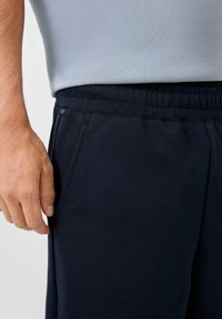 Close-up van een persoon die een marineblauwe elastische broek met zijzakken en een lichtgrijze top draagt.