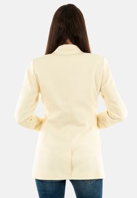 Blazer color crema con un corte ajustado, mangas largas, cuello con muesca y una textura suave. Visto desde la parte posterior, combinado con vaqueros.