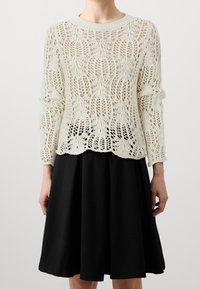 Kremowy sweter na drutach z koronkowym wzorem i okrągłym dekoltem, zestawiony z czarną rozkloszowaną spódnicą o długości do połowy łydki i gładkiej fakturze.