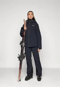 Tumši zils slēpošanas jaka un bikses no ūdensnecaurlaidīga auduma, ar rāvējslēdzēja aizdari, regulējamu kapuci un melniem slēpju zābakiem, kas tur slēpji.