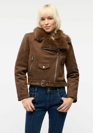 Giovane donna con capelli biondi che indossa una giacca di camoscio marrone con colletto in pelliccia e jeans blu, in piedi con le mani nelle tasche.