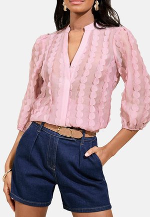 Blouse rose transparente avec un motif circulaire, manches bouffantes longues et boutonnage à l'avant. Associée à un short en denim bleu foncé et une ceinture beige.