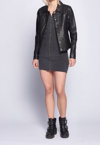 Zwarte leren jas over een donkergrijze aansluitende jurk met een rits aan de voorkant. Zwarte gestudeerde enkellaarsjes, voorzien van gespen en dikke zolen.