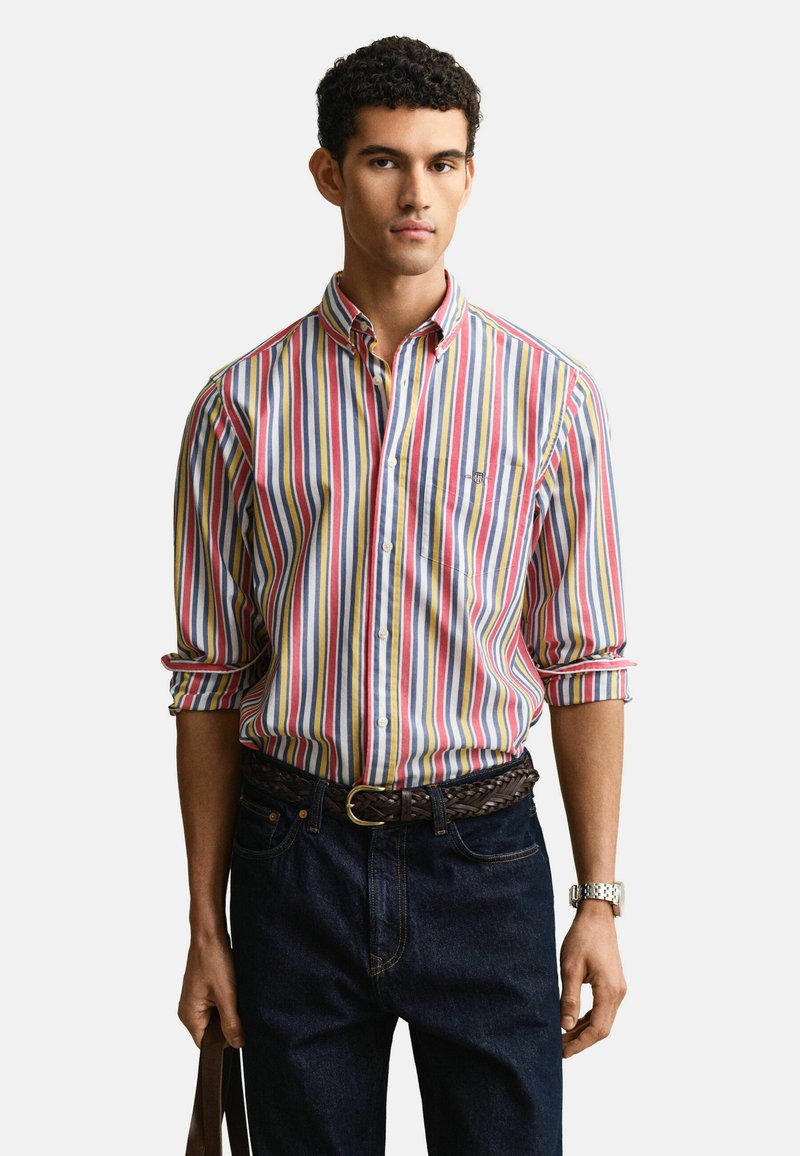 Jeune homme portant une chemise à rayures verticales multicolores avec manches retroussées, jean foncé, ceinture tressée marron, montre-bracelet argentée, tenant un sac marron.